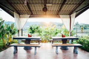 An outdoor seating area with two wooden tables and chairs, surrounded by lush greenery and a view of distant hills.