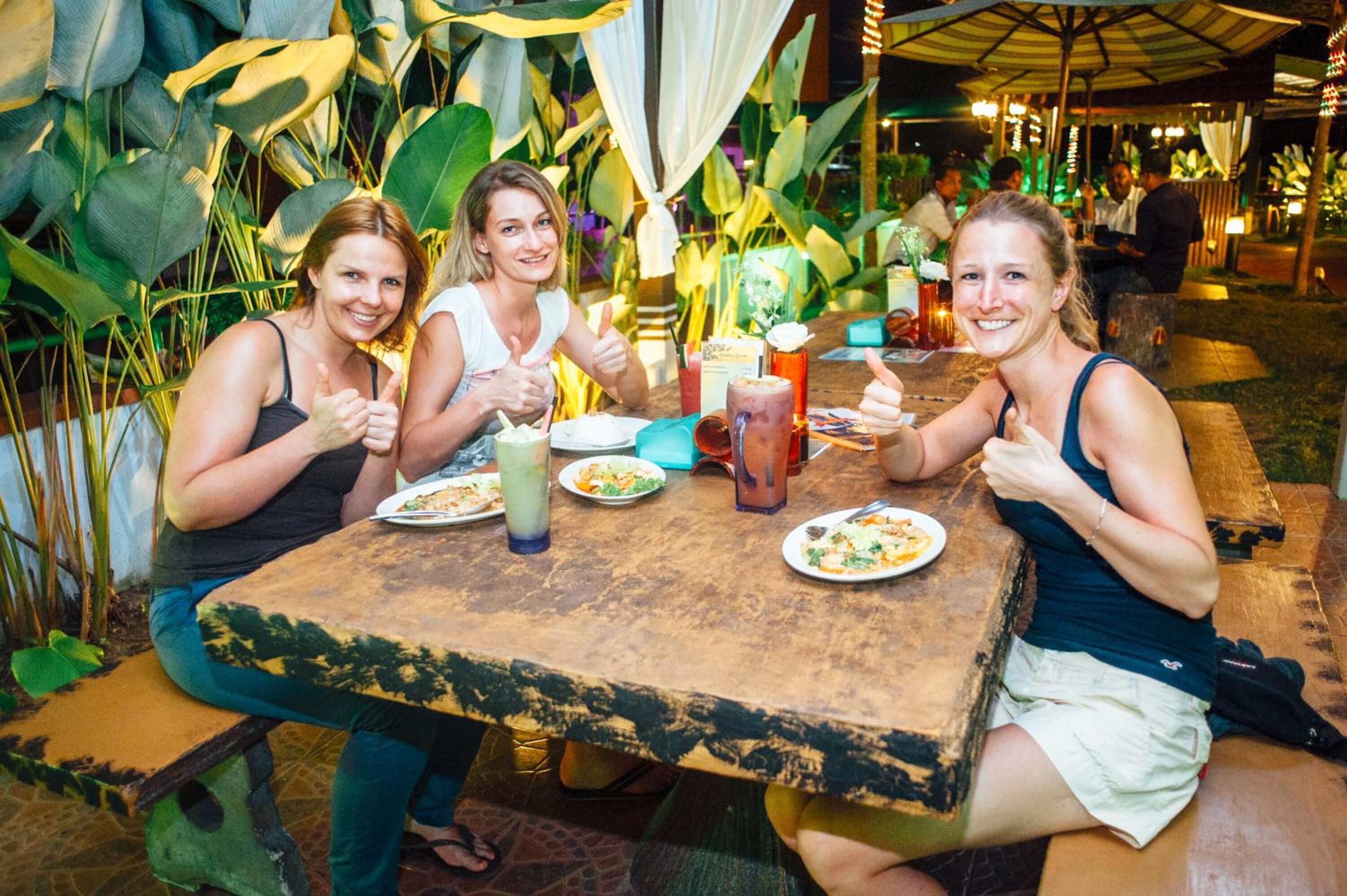 A group of people dining at an outdoor table with a rustic wooden table and a lush green backdrop.