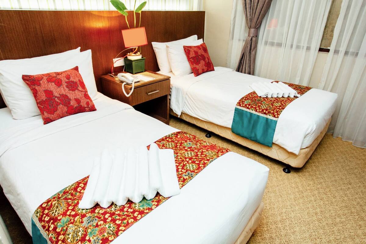 A hotel room with two beds, each with white linens and red floral pillows. There is a nightstand with a lamp and a telephone. The room has a wooden headboard and a window with sheer curtains.