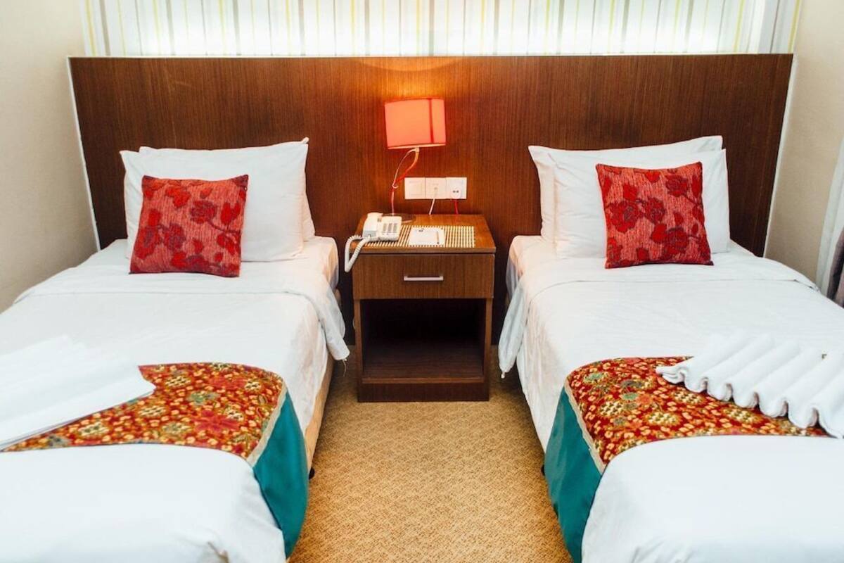A hotel room with two beds, each with a red patterned pillow and a white bedspread. There is a nightstand between the beds with a lamp and a phone.