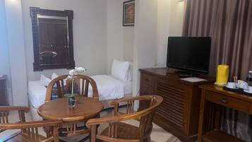 A hotel room with a wooden dining table, four chairs, a sofa, a TV on a wooden cabinet, and a bed with white bedding.