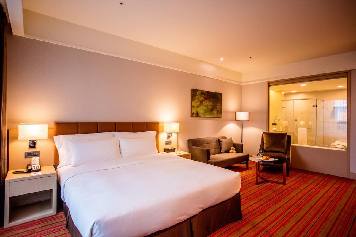 A hotel room with a large bed, two bedside tables with lamps, a chair, a small table with a plate of fruit, and a painting on the wall.