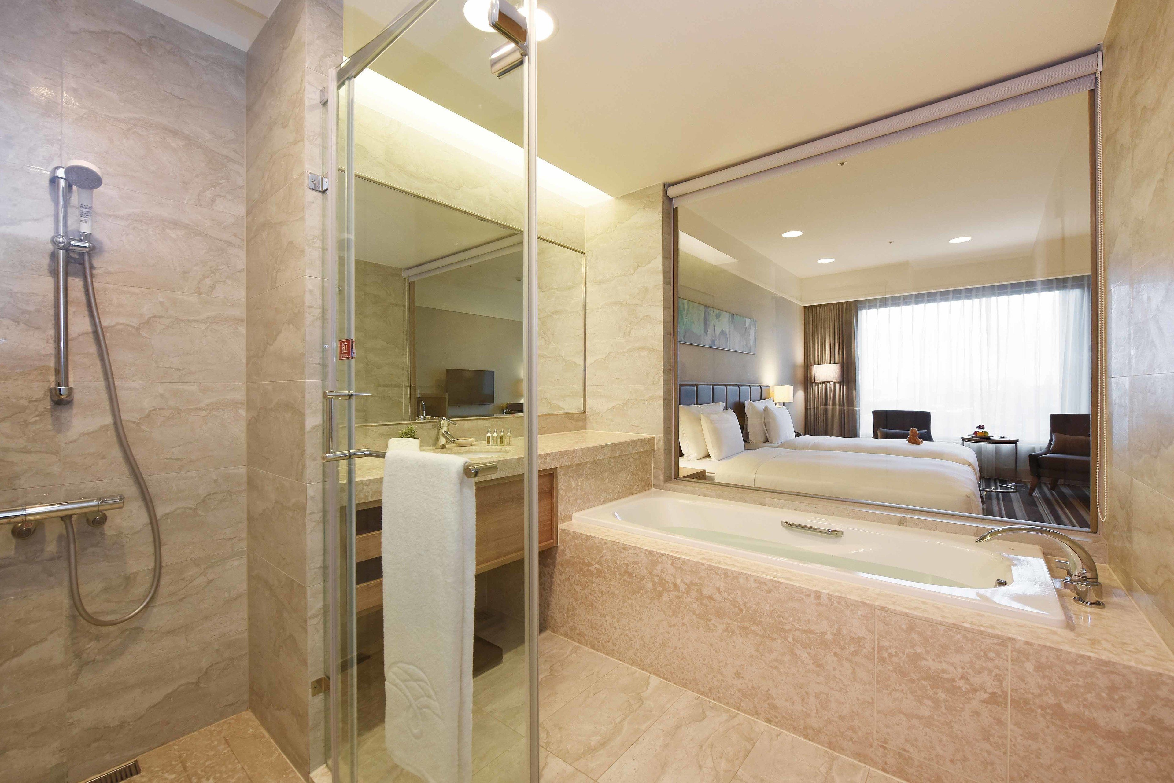 Un baño moderno con ducha envuelta en vidrio, un espejo grande, una bañera y una habitación de hotel visible con cama y escritorio.