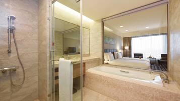 Un baño moderno con ducha envuelta en vidrio, un espejo grande, una bañera y una habitación de hotel visible con cama y escritorio.