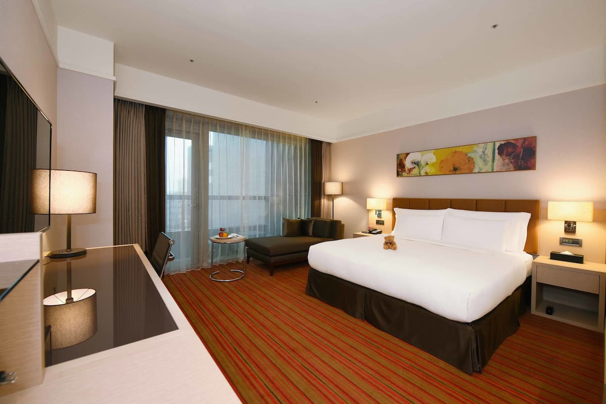 A hotel room with a large bed, a desk, a chair, a small table, and a large window with curtains.