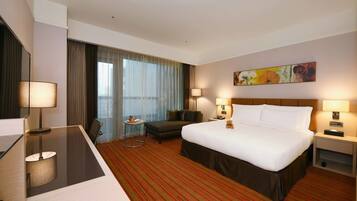 A hotel room with a large bed, a desk, a chair, a small table, and a large window with curtains.
