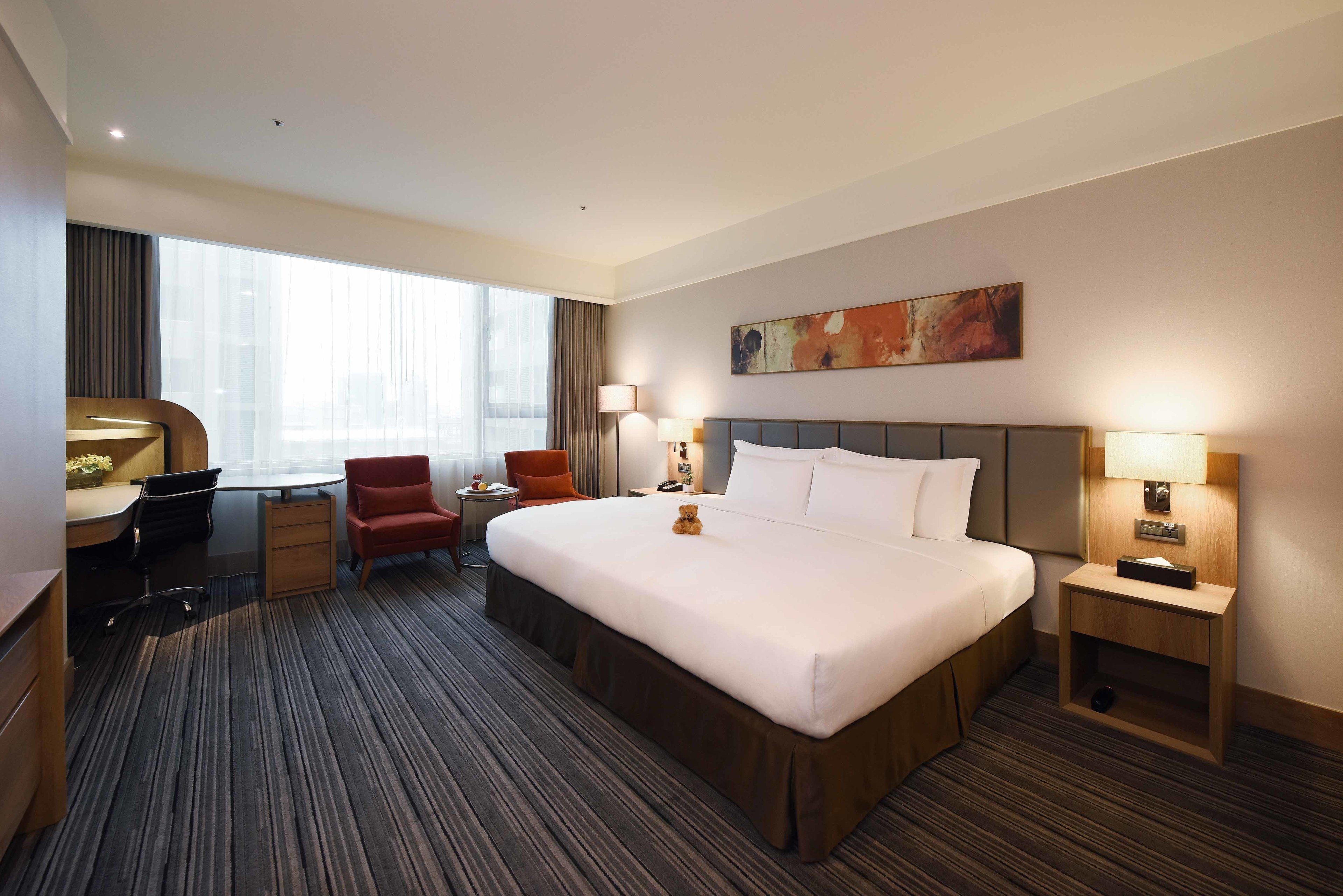 A hotel room with a large bed, a desk with a chair, a small table with a lamp, and a large window with curtains.
