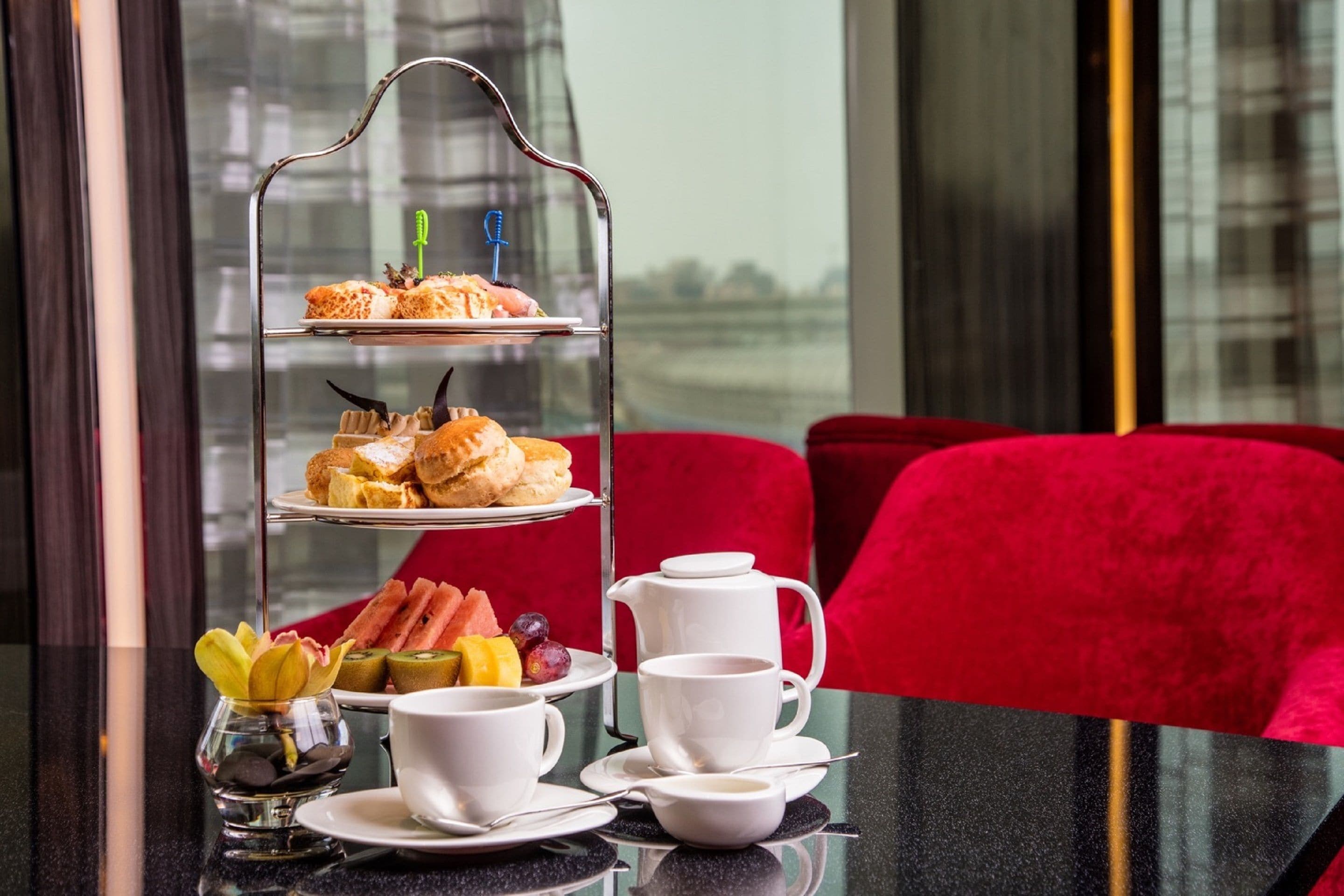 A three-tiered dessert stand with pastries and fruits, white teacups, and a small vase with flowers on a table.