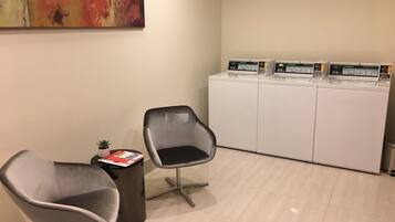 A reception area with two grey chairs, a small table with a magazine and a plant, and a row of white washing machines with digital displays.