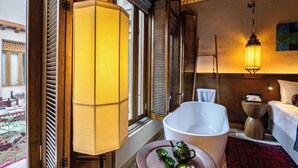 Une pièce avec une grande fenêtre, une table de chevet surmontée d’une lampe blanche, une commode en bois et un tapis rouge.