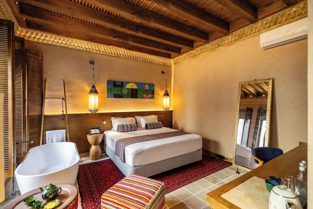Ein Hotelzimmer mit einem großen Bett, Holzbalkendecke, rotem Teppich, freistehender weißer Badewanne, farbigem Pouf und einem Spiegel mit goldener Rahmung.