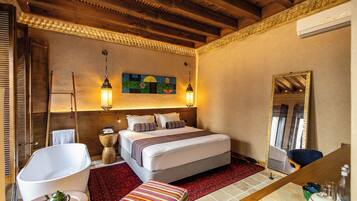 A hotel room with a large bed, a wooden ceiling, a red carpet, a white bathtub, a colorful ottoman, and a mirror with a gold frame.