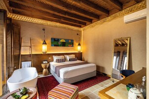 A hotel room with a large bed, a wooden ceiling, a red carpet, a white bathtub, a colorful ottoman, and a mirror with a gold frame.