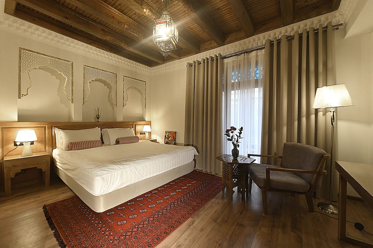 Ein Hotelzimmer mit einem großen Bett, einem Holzkopfende, zwei Nachttischlampen, einem roten Teppich mit Muster, einem Holznachttisch mit einer Vase, einem Sessel und einem großen Fenster mit Vorhängen.