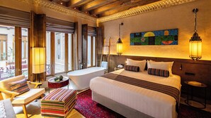 Une chambre d’hôtel spacieuse, dotée d’un grand lit, d’un coin salon avec un fauteuil et une petite table, d’une baignoire et d’une grande fenêtre donnant sur l’extérieur.