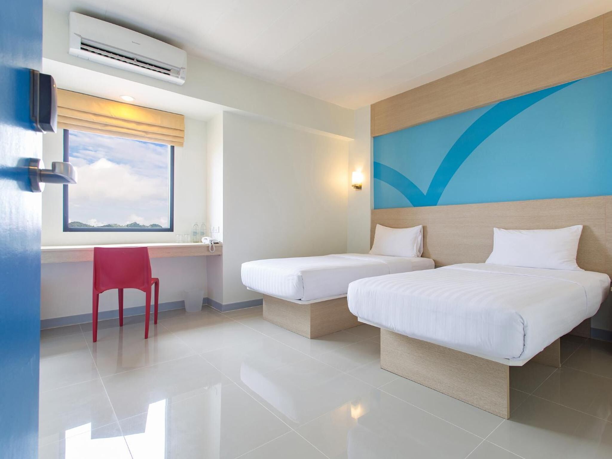 A hotel room with two beds, a desk, a chair, an air conditioning unit, and a window with a view.