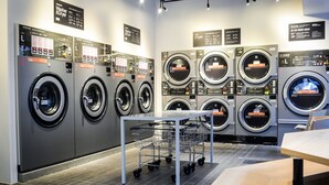 Une laverie équipée de plusieurs machines à laver et de plusieurs sèche-linge, d’un chariot en métal et d’une table.