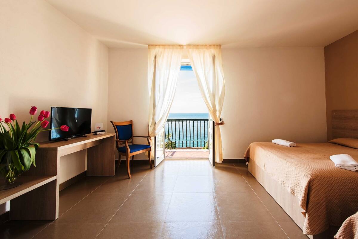 Ein Hotelzimmer mit Bett, Schreibtisch mit Fernseher, Stuhl und Balkon mit Meerblick.