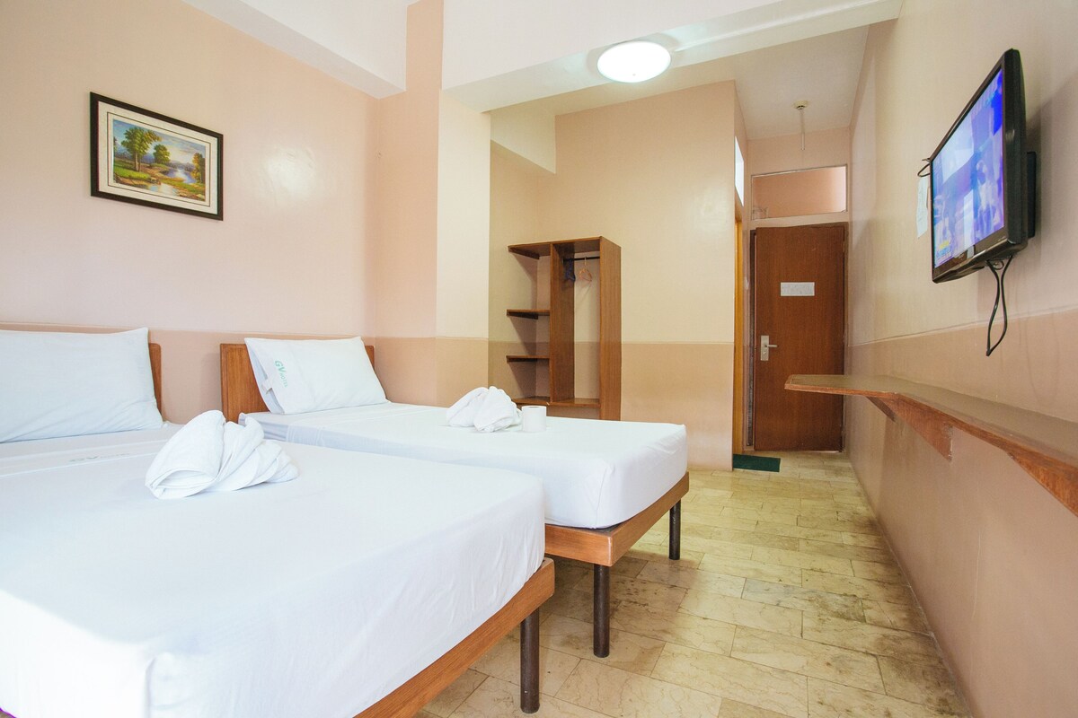 Una habitación de hotel con dos camas individuales, un armario de madera, un televisor montado en la pared y un cuadro.