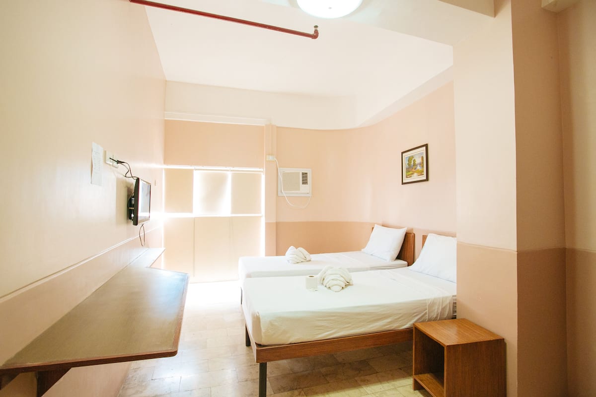 A hotel room with two beds, a wooden bench, a small wooden side table, a wall-mounted TV, and a picture on the wall.
