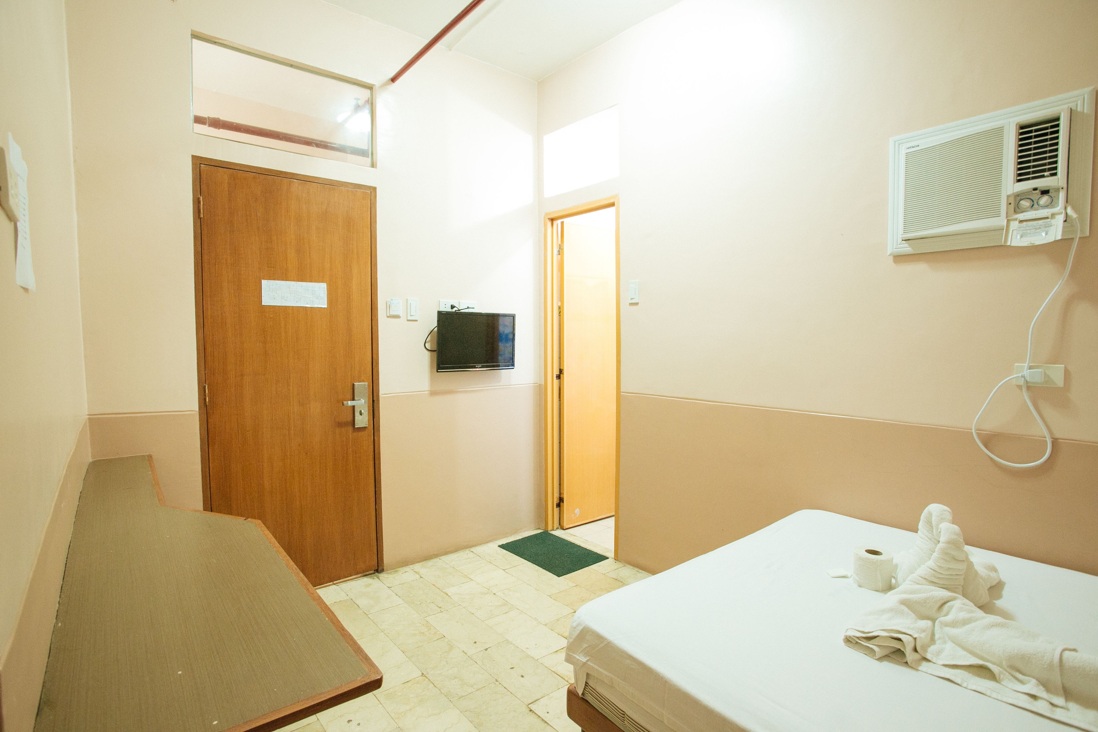 A hotel room with a bed, a wooden desk, a television, and a wall-mounted air conditioner.