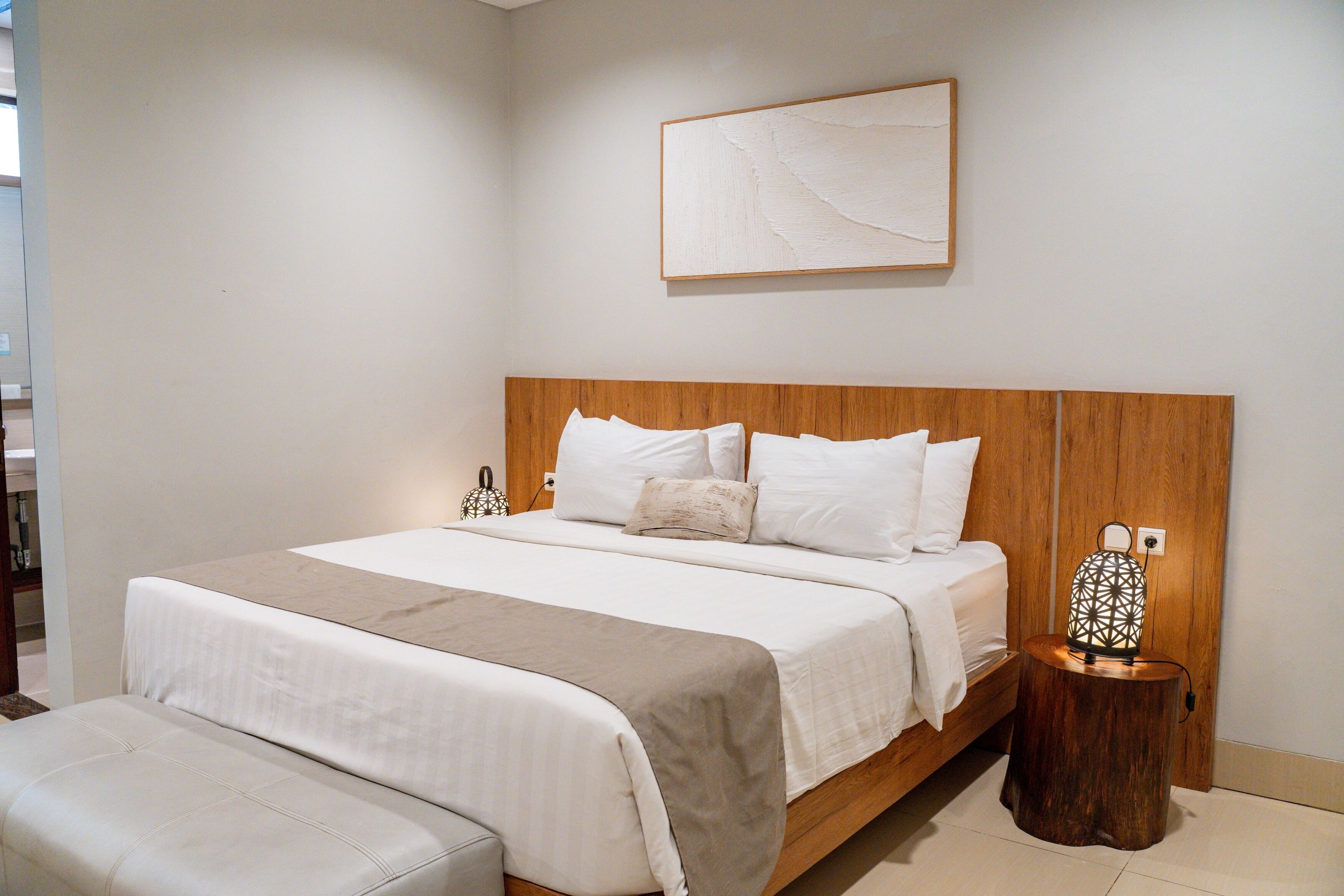 Une chambre à coucher avec une tête de lit en bois, un grand lit avec une literie blanche, une table de chevet ornée d’une lanterne décorative et un tableau abstrait accroché au mur.