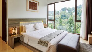 Un cuarto de hotel con una cama grande, una mesita de noche con lámpara, un cuadro enmarcado en la pared y un ventanal con vista a follaje.