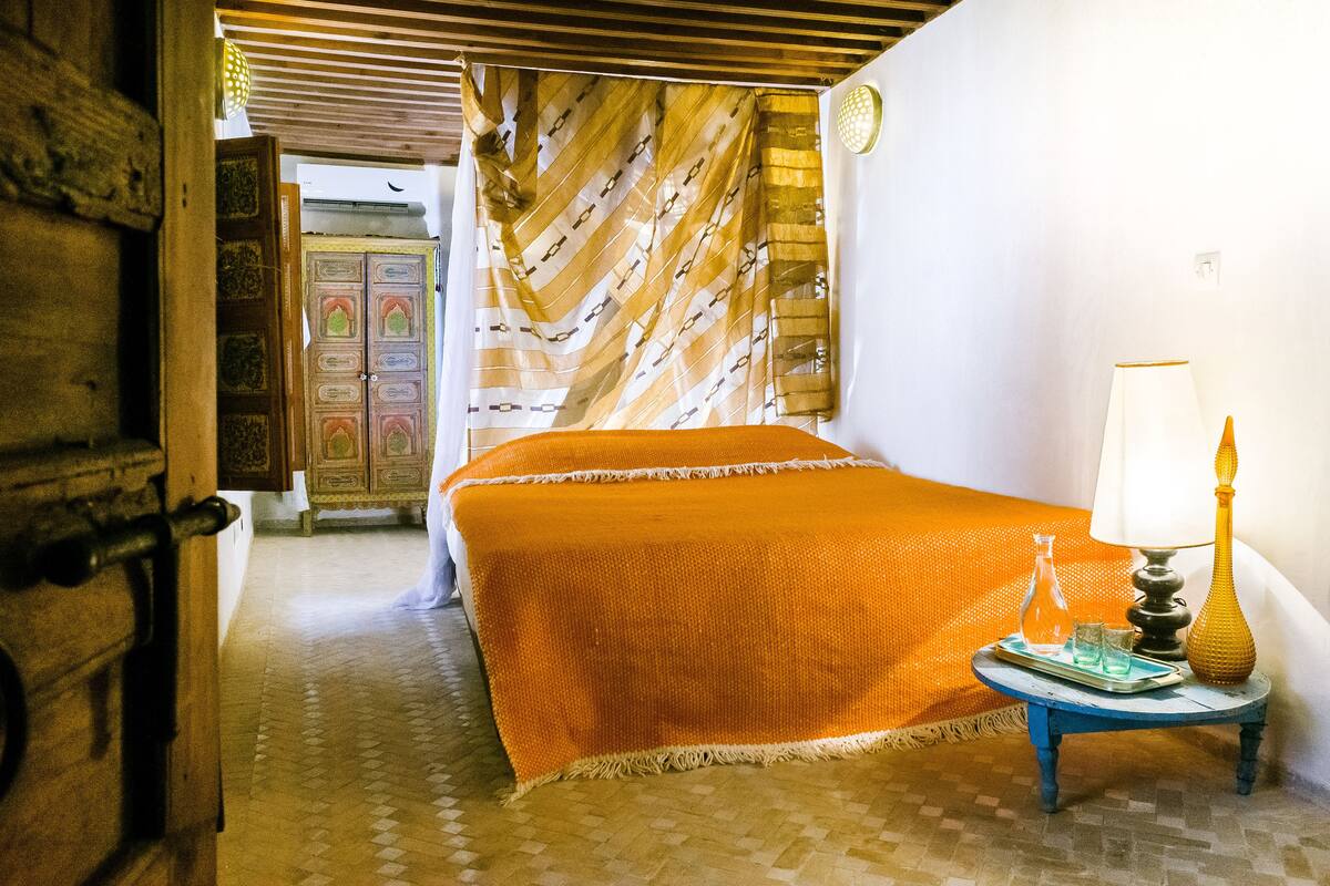 A room with a bed, a nightstand with a lamp, a patterned curtain, and a door leading to another room.