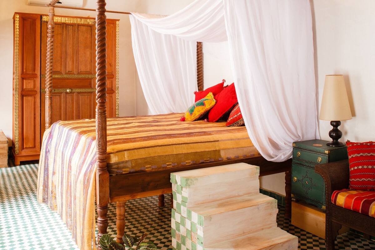 A four-poster bed with white drapes, a wooden floor with a checkered pattern, and a green plant in a square planter.
