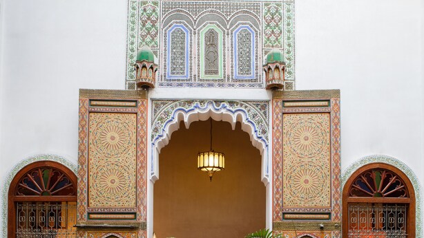 An ornate Moroccan interior with a red carpet, a large brass pot, and two potted plants.