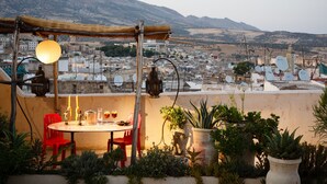 A rooftop terrace with a table set for dining, surrounded by potted plants and a view of a cityscape with mountains in the background.