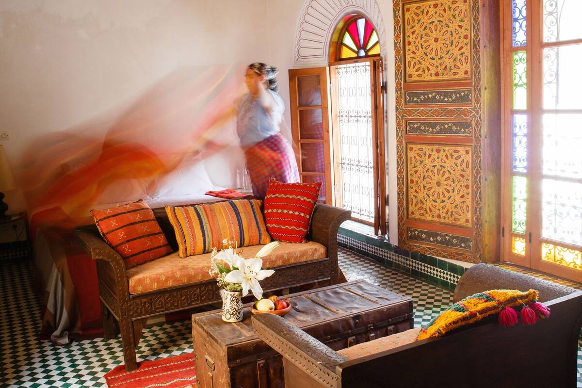 A traditional Moroccan interior with a checkered floor, a sofa with colorful cushions, a wooden coffee table with a vase and fruit bowl, and a decorative door with intricate patterns.