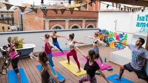 Un grupo de personas practicando yoga en una terraza de madera, con esterillas de colores, un mural grande y un edificio de ladrillo al fondo.