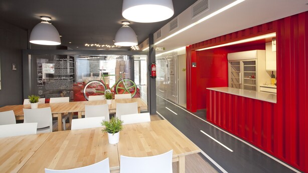 Un comedor moderno con mesas de madera y sillas blancas, una pared de acento roja y una bicicleta al fondo.