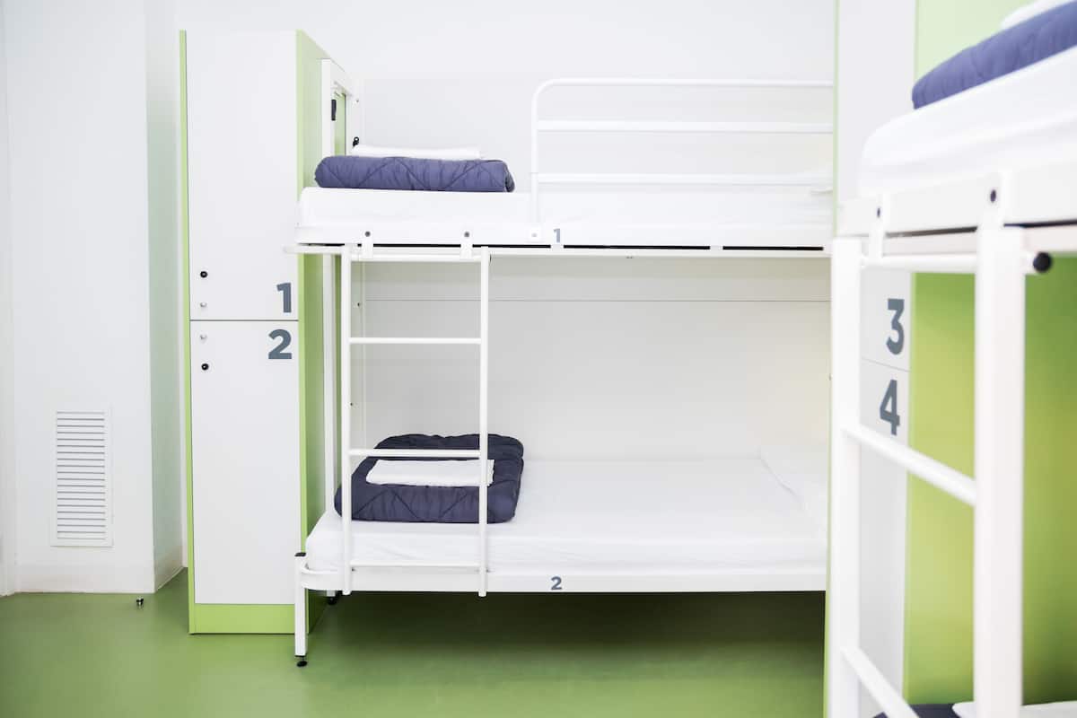 Habitación con literas numeradas para cuatro personas, piso verde y pared blanca con una rejilla de ventilación.
