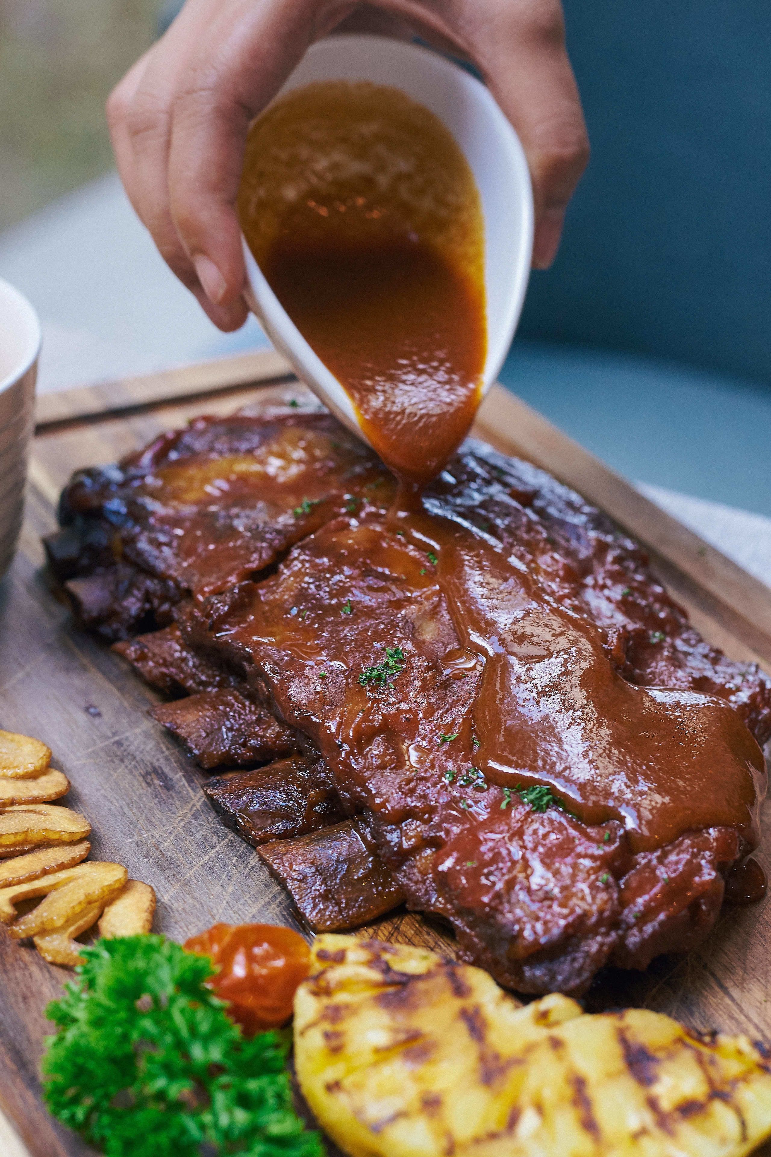 Una persona sirviendo salsa sobre un plato de costillas en una tabla de madera.