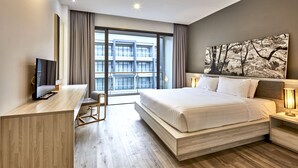 A modern hotel room with a large bed, a desk, a television, and a view of the cityscape through large windows. - Serenity Hotel Spa & Onsen (Kabin Buri)