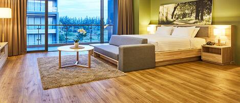 A modern hotel room with a large bed, a sofa, a small table, and a nightstand. There is a large window with a view of trees and a building.