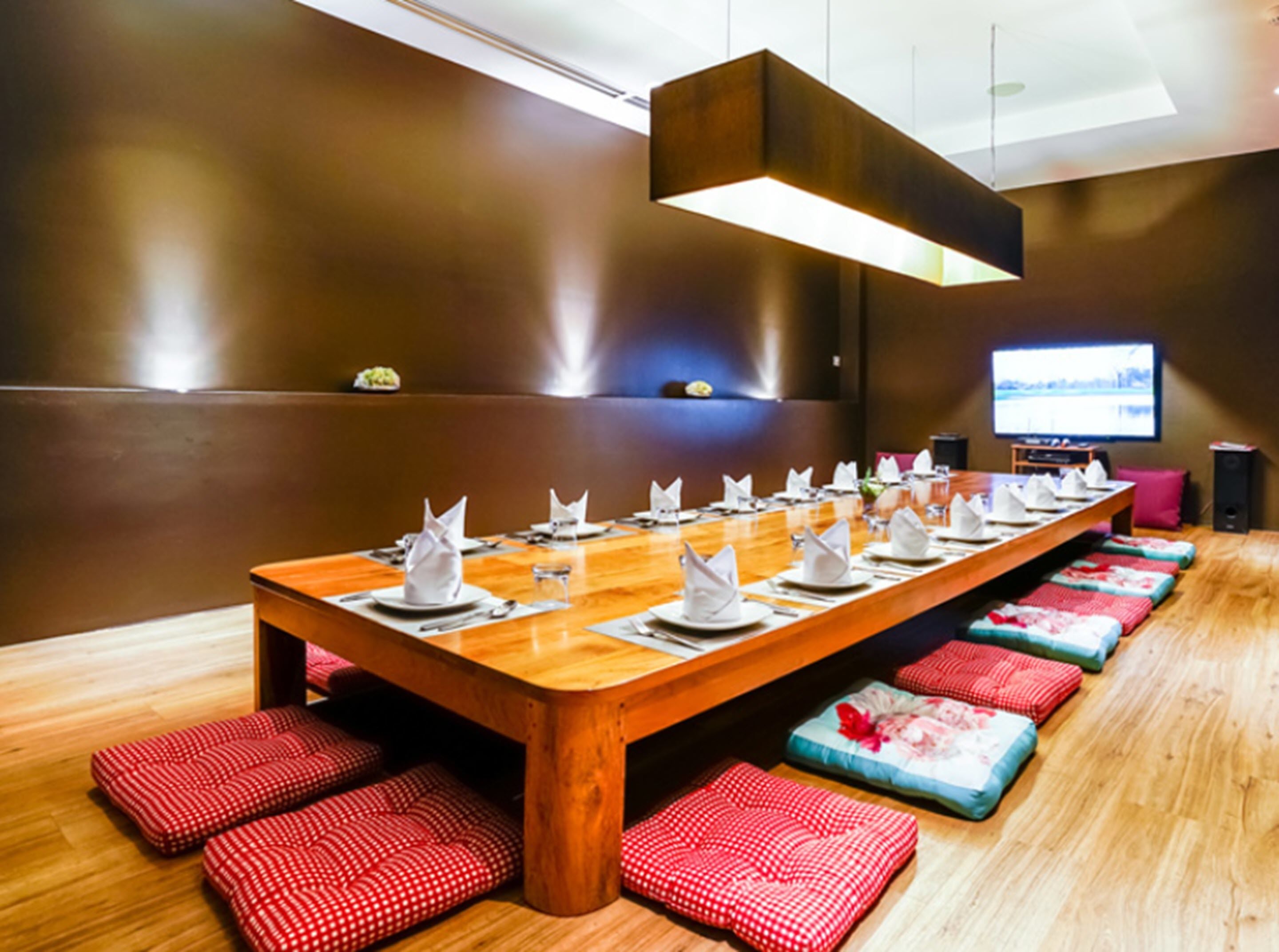 A long wooden dining table set with white napkins, plates, and glasses, surrounded by red and blue cushioned seating.