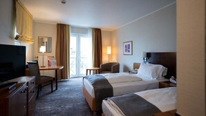 A hotel room with two beds, a desk with a chair, a television, and a large window with curtains.