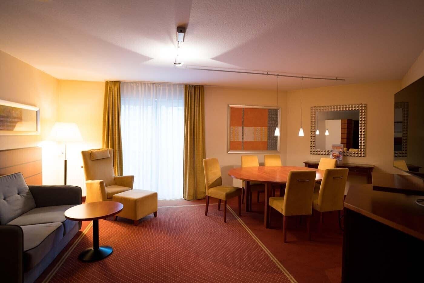 Habitación de hotel con sofá, sillón, mesa de comedor, sillas, una mesita redonda, lámpara, espejo y ventana con cortinas.