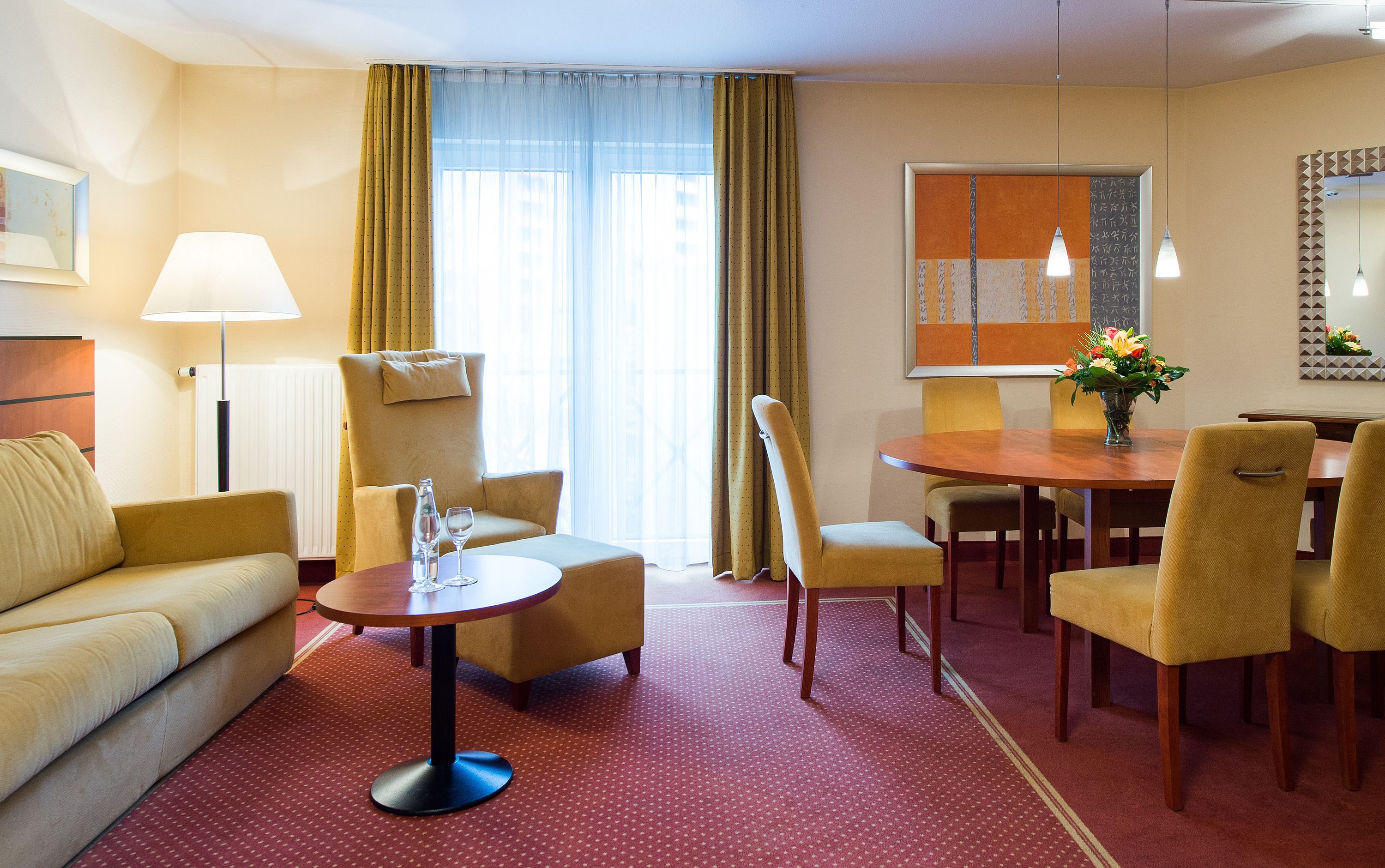 Una habitación de hotel con un sofá, una mesa de comedor redonda con sillas, una lámpara y una ventana con cortinas.
