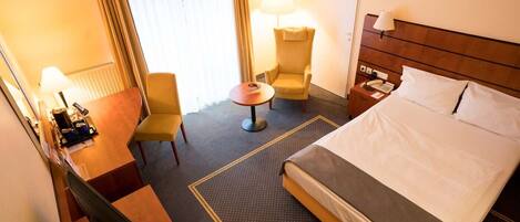 A hotel room with a bed, a desk, a chair, a small table, and a lamp.