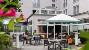 An outdoor patio area with tables and chairs, a covered seating area, and a building with large windows and balconies. - Hotel Fulda Mitte (Fulda)