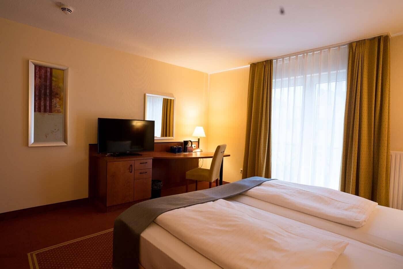 Una habitación de hotel con una cama, un escritorio con televisor, una silla y una ventana con cortinas.