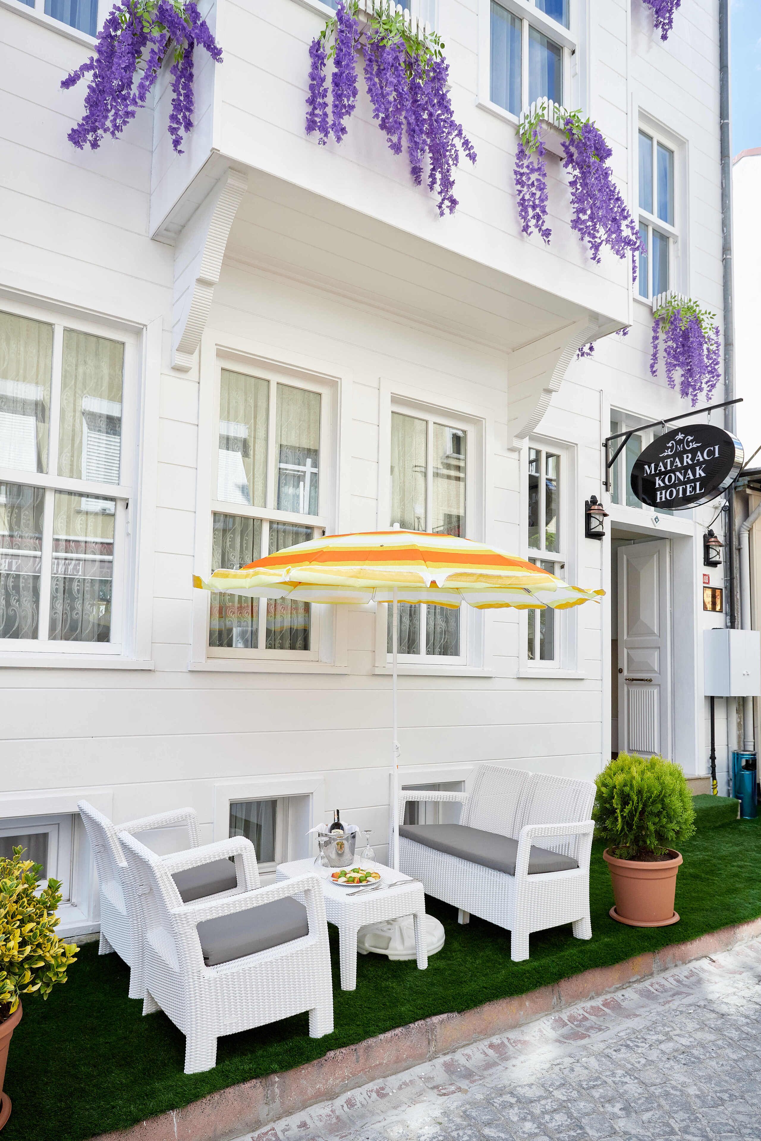 A hotel exterior with white walls, purple hanging plants, and outdoor seating with white wicker furniture.
