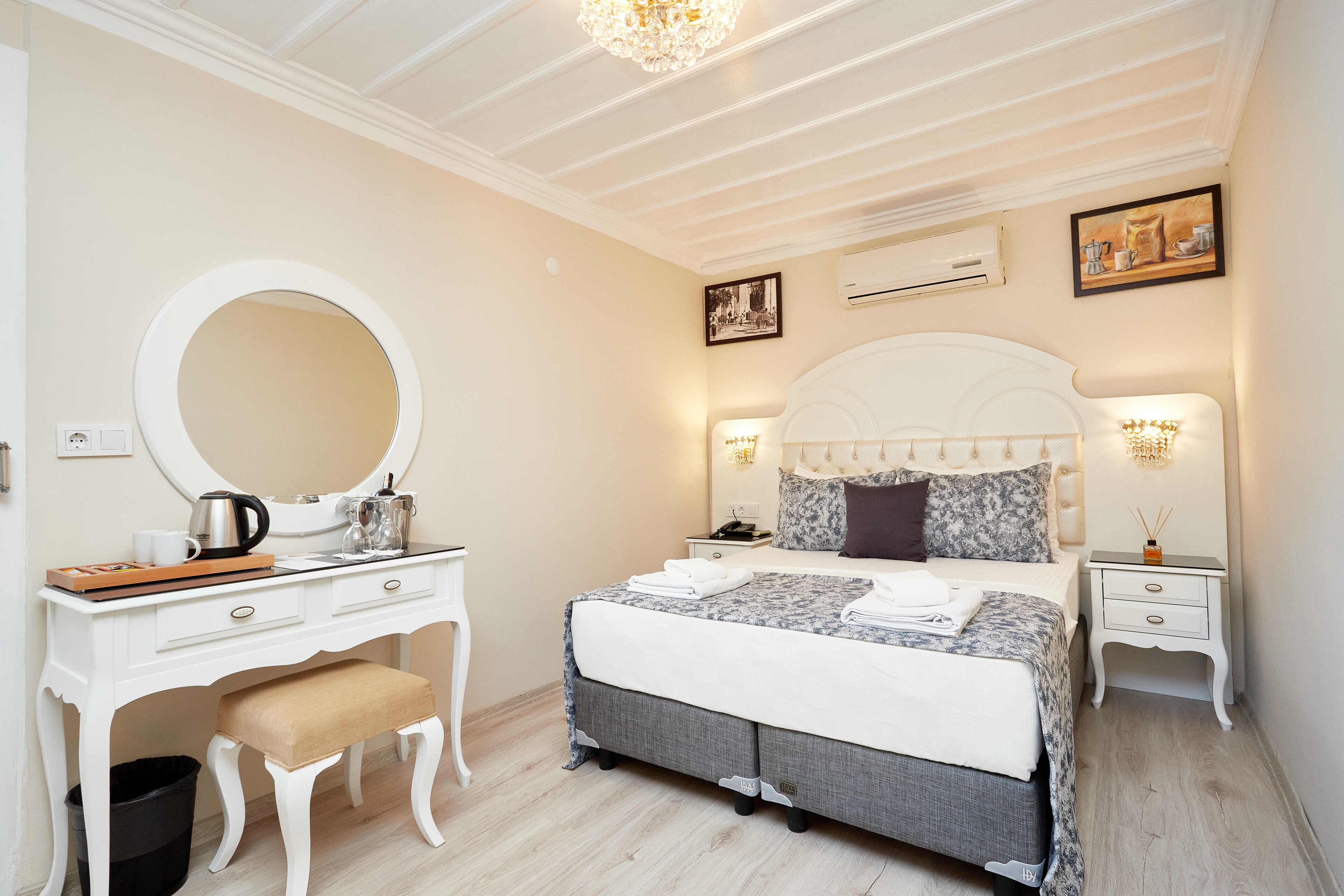 A bedroom with a white vanity, a large round mirror, a bed with a grey and white duvet, a white headboard, and two bedside tables with lamps.