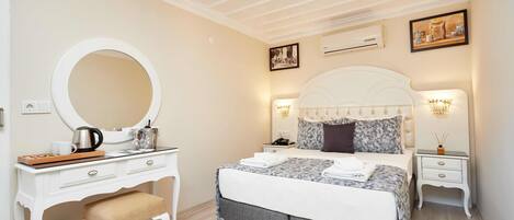 A bedroom with a white vanity, a large round mirror, a bed with a grey and white duvet, a white headboard, and two bedside tables with lamps.