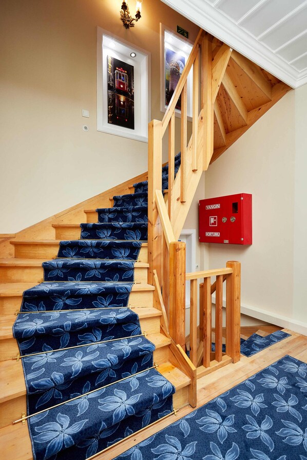 Un escalier en bois avec des marches recouvertes d’une moquette bleue et une boîte d’alarme incendie rouge fixée au mur.