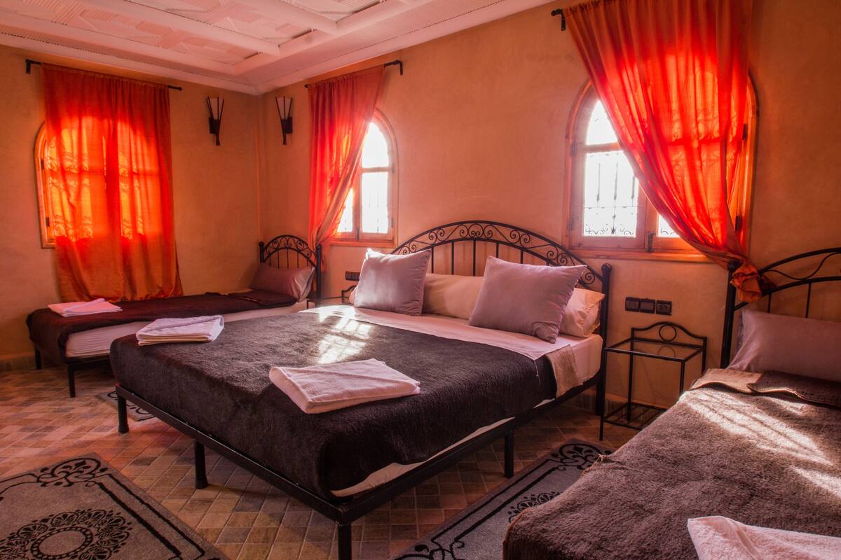 A bedroom with a large bed, two smaller beds, and a bench. There are curtains on the windows, and the room has a tiled floor.